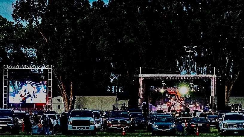 people outside their cars attending a drive-in concert featuring a mobile stage and LED screen on a truss structure